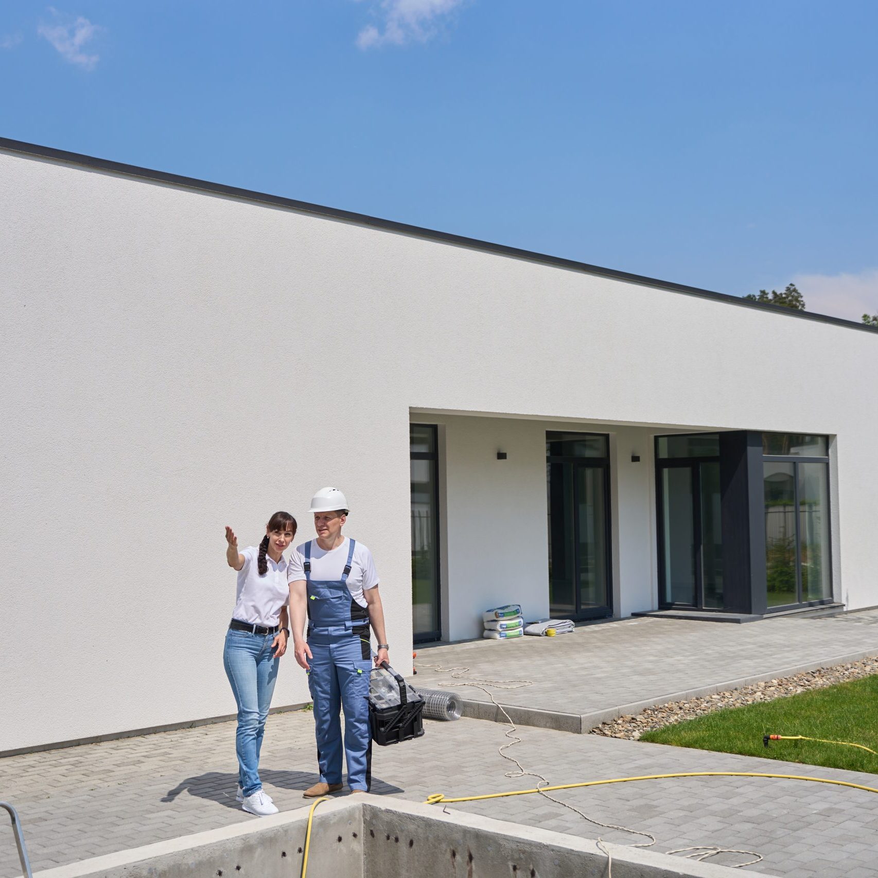 Woman showing something to repairman while they standing by empty swimming pool before repair near new modern townhouse in warm summer day